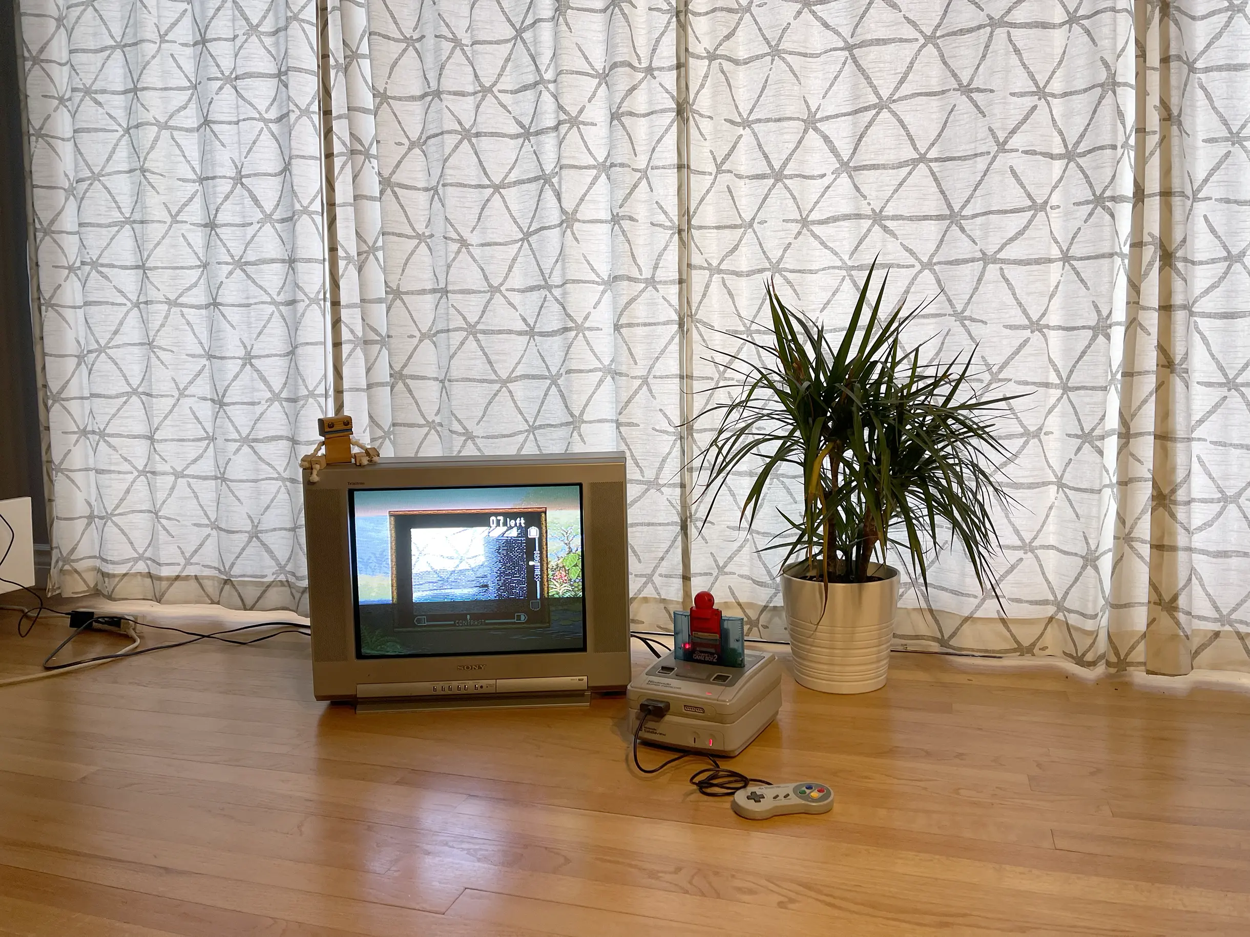 A Japanese Super Famicom console with Super Game Boy adapter, Game Boy Camera, Bandai Satellaview, staged beside a Sony Trinitron CRT, potted plant, and wooden robot.