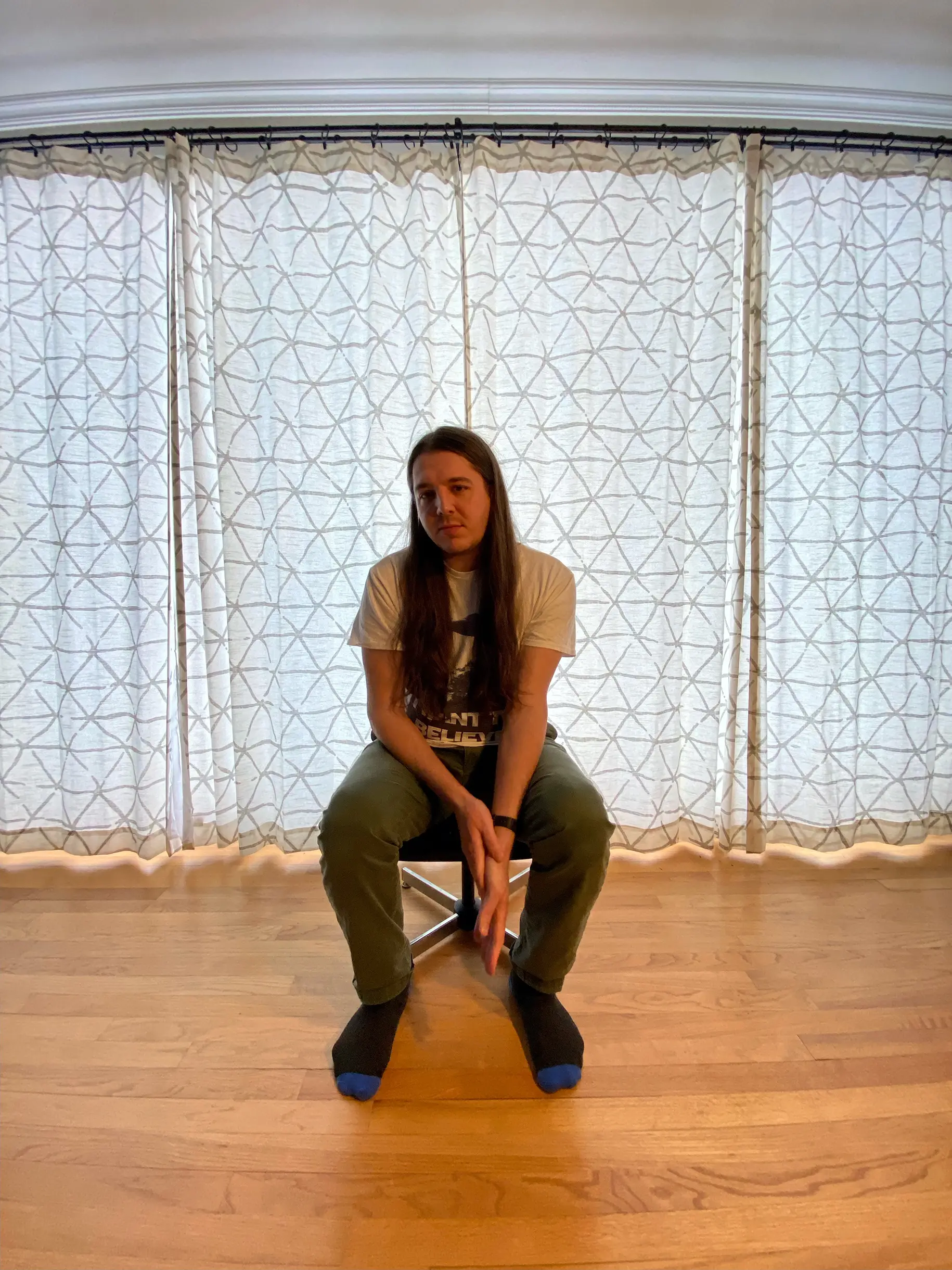 James Edward, musician, sits in front of drawn curtains on a vintage footrest in a behind-the-scenes photo from the Desire music video shoot.